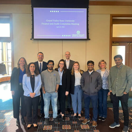 College of computing's faculty and staff stand together and smiles for a photo in front of a screen displaying “Grand Valley State University Finance and Audit Committee Meeting – April 25, 2025.”
